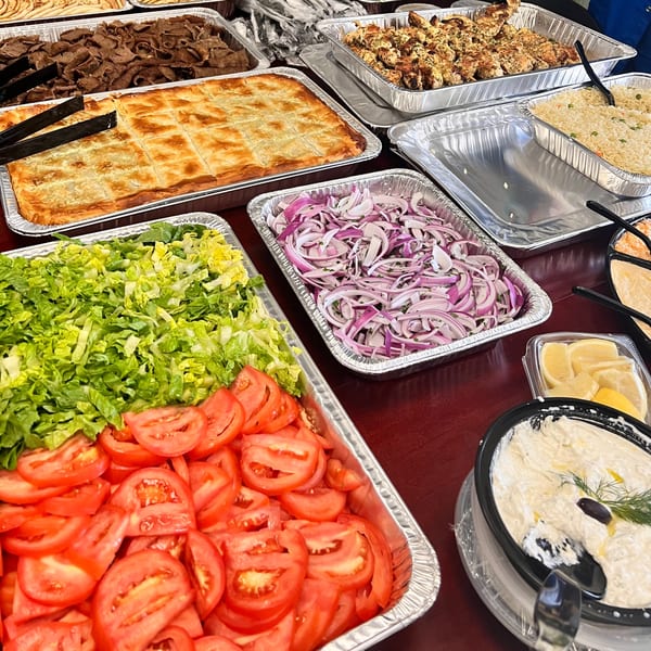 A Greek catering spread with gyro meat, spanakopita, chicken skewers, rice, salad, and tzatziki.