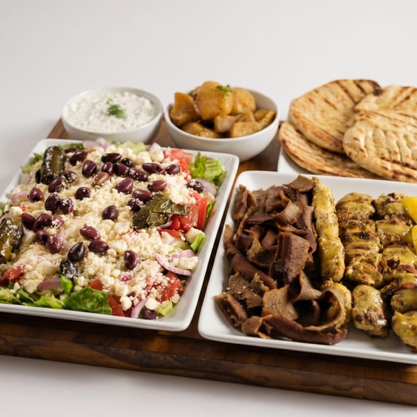 Greek dinner with gyro meat, chicken skewers, Greek salad, roasted potatoes, tzatziki, and pita bread.