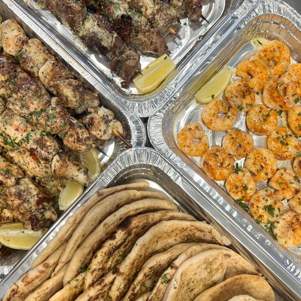 Catering trays with chicken and beef skewers, seasoned shrimp, and pita bread.