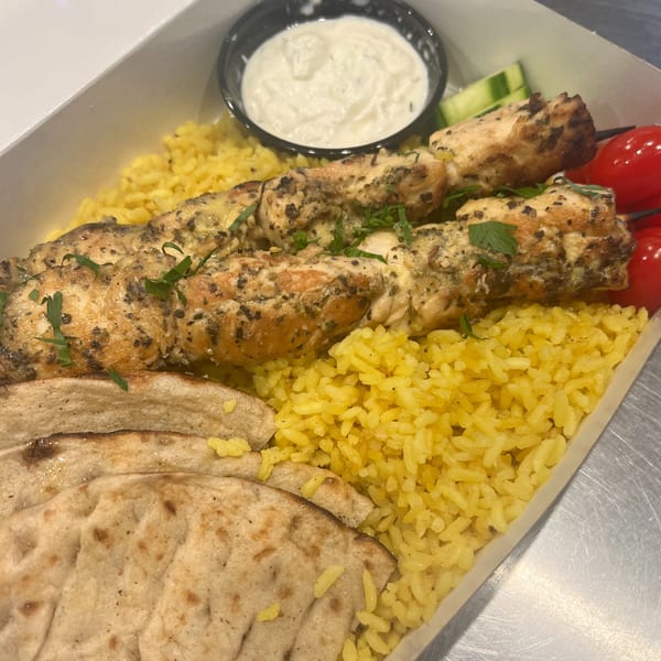 Chicken souvlaki skewers with yellow rice, pita bread, tzatziki sauce, and cherry tomatoes in a takeout container.