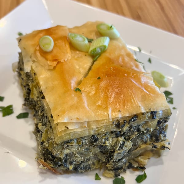 A slice of golden-brown Greek spanakopita, garnished with fresh green onions, on a white plate.
