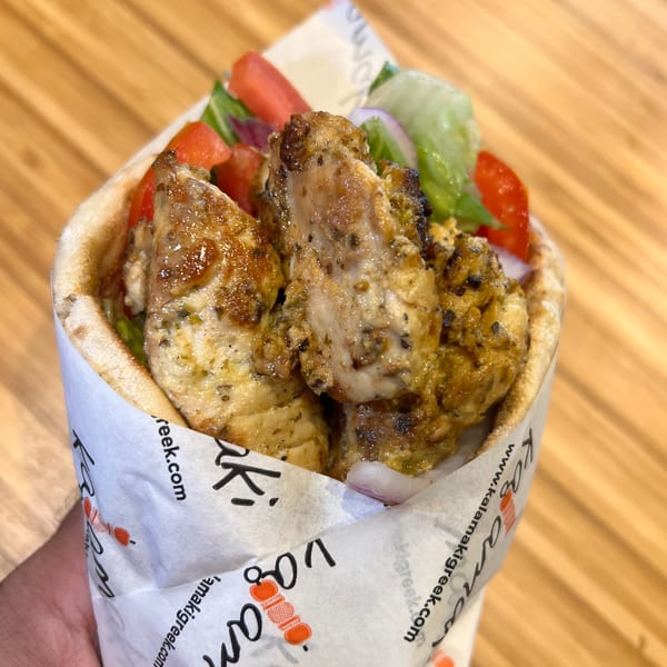 Hand holding a Kalamaki Greek chicken souvlaki pita wrap with tomatoes, lettuce, and onions.
